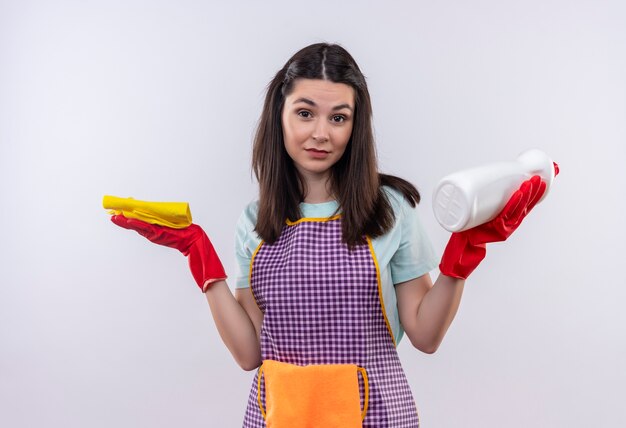 Young beautiful girl in apron and rubber gloves holding cleaning supplies and sponge looking confused shrugging shoulders 