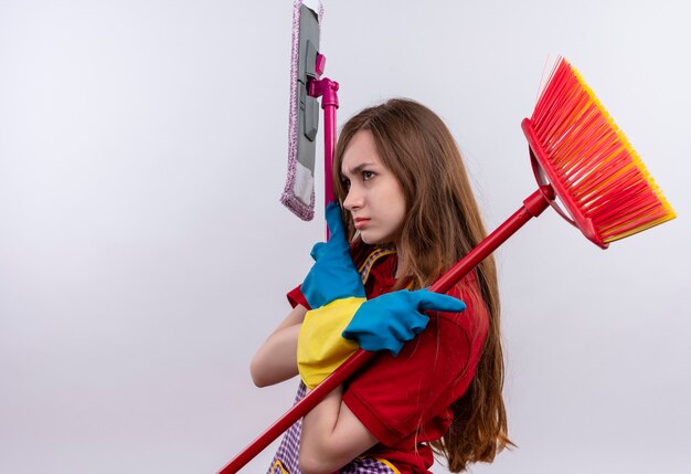 Young beautiful girl in apron and rubber gloves  holding mops frowning, sideways 