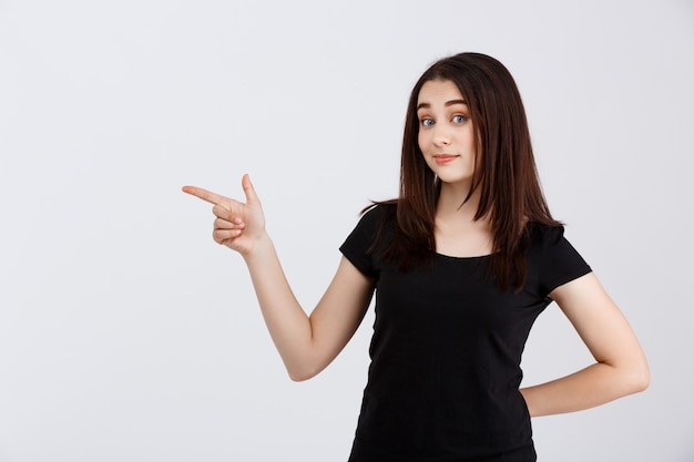 Young beautiful girl in black t-shirt pointing fingers in side over white wall