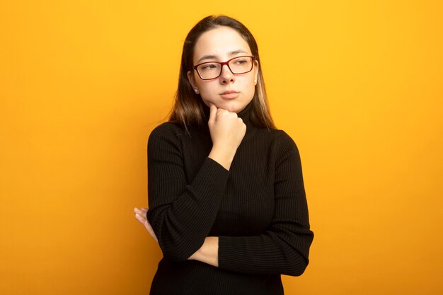 Young beautiful girl in a black turtleneck looking aside with hand on chin thinking 