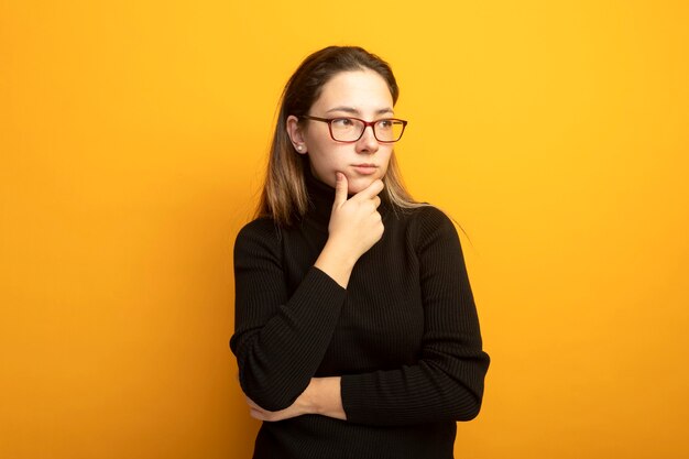 Young beautiful girl in a black turtleneck looking aside with hand on her chin thinking 