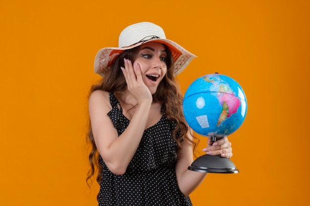 Free Photo young beautiful girl in dress in polka dot in summer hat holding globe looking at it amazed and surprised standing over yellow background