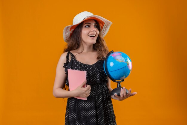 Young beautiful girl in dress in polka dot in summer hat holding globe and notebook looking aside smiling with happy face standing over yellow background