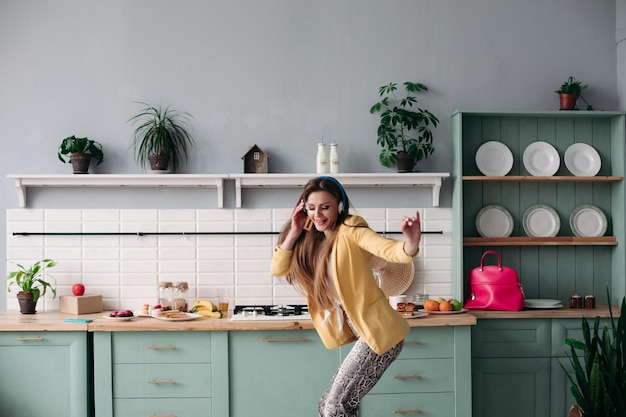 Free photo young beautiful girl in fashionable clothes getting fun on kitchen happy brunette model with long hair listening to music and dancing stylish woman in yellow jacket and blue headphones singing