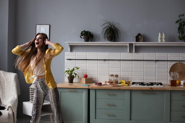 Young beautiful girl in fashionable clothes getting fun on kitchen Happy brunette model with long hair listening to music and dancing Stylish woman in yellow jacket and blue headphones singing