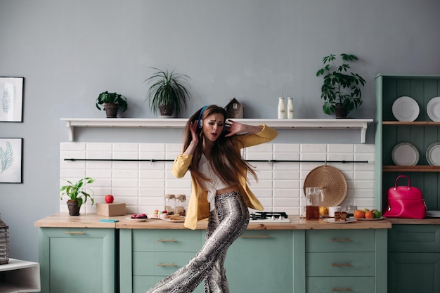 Free photo young beautiful girl in fashionable clothes getting fun on kitchen happy brunette model with long hair listening to music and dancing stylish woman in yellow jacket and blue headphones singing