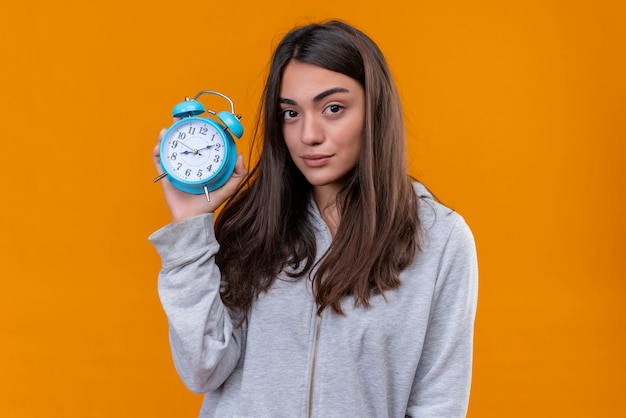 Young beautiful girl in gray hoody holding clock and looking at camera with pensive expression standing over orange background