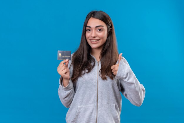 Young beautiful girl in gray hoody holding credit card and looking at camera with smile on face and make like gesture standing over blue background