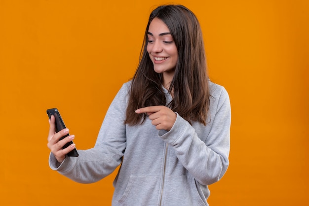 Young beautiful girl in gray hoody holding telephone and pointing to telephone  at the same time looking to telephone with smile on face standing over orange background