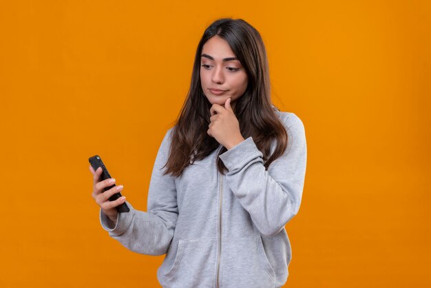 Young beautiful girl in gray hoody holding telephone standing over orange background   looking to telephone with pensive sight standing