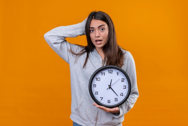 Young beautiful girl in gray hoody look at camera standing with hand on head for mistake and other hand with clock on her hand standing over orange background