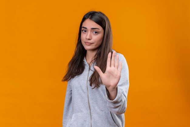 Young beautiful girl in gray hoody looking at camera weary sight on face and making stop gesture standing over orange background