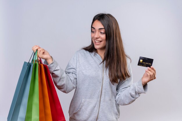 Young beautiful girl in gray hoody looking to packages with smile on face holding packages and credit card standing over white background  white background