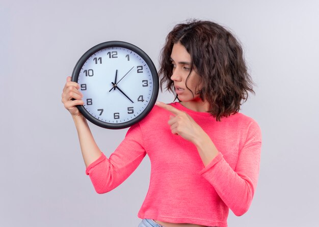 Young beautiful girl holding clock and pointing at it on isolated white background