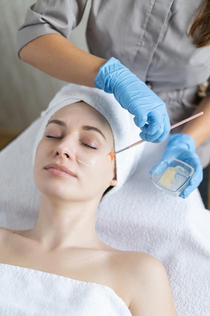 A young beautiful girl lies on the table at the beautician, hands in blue gloves apply a moisturizing cream, a mask with a brush