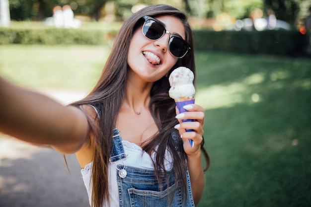 Young beautiful girl makes selfie smiles with white teeth and holds the ice cream wearing sunglasses