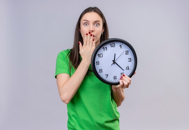 Young beautiful girl wearing green t-shirt holding clock looking surprised and shocked covering mouth with hand standing over isolated white background