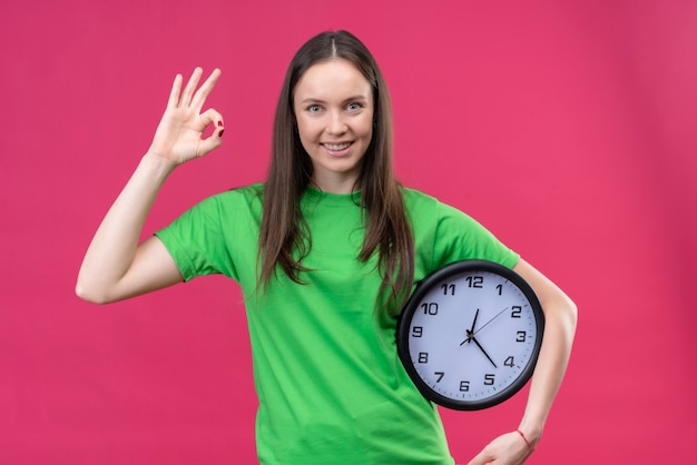 Free photo young beautiful girl wearing green t-shirt holding clock smiling cheerfully doing ok sign standing over isolated pink background