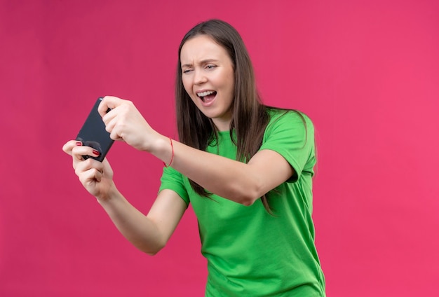 Young beautiful girl wearing green t-shirt holding smartphone looking at it stressed and nervous standing over isolated pink background