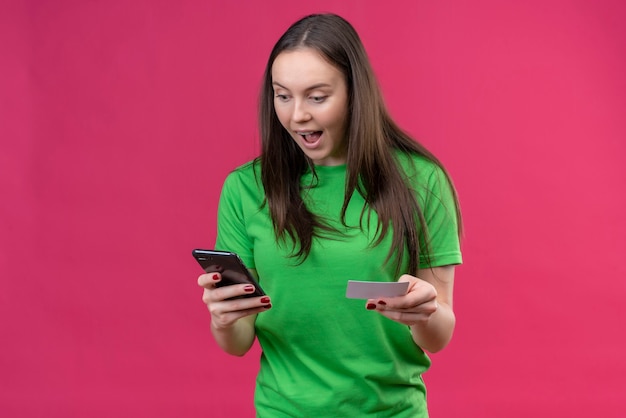 Young beautiful girl wearing green t-shirt holding smartphone looking at screen amazed and surprised standing over isolated pink background