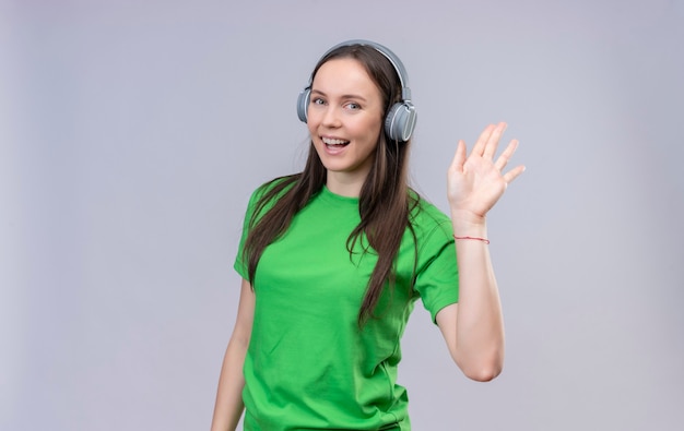 Young beautiful girl wearing green t-shirt with headphones smiling cheerfully waving with hand standing over isolated white background