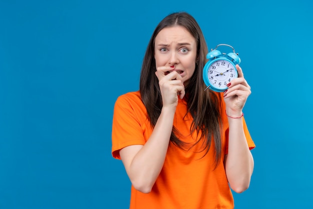 Free photo young beautiful girl wearing orange t-shirt holding alarm clock stressed and nervous biting her nails standing over isolated blue background