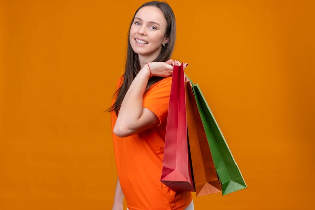 Young beautiful girl wearing orange t-shirt holding paper bags looking at camera smiling cheerfully standing over isolated orange background