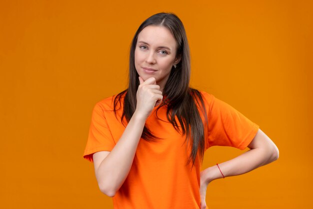 Young beautiful girl wearing orange t-shirt looking at camera with hand on chin thinking standing over isolated orange background