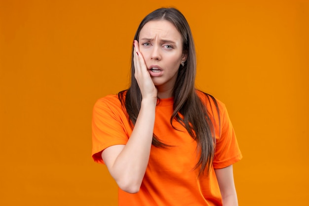 Young beautiful girl wearing orange t-shirt looking unwell touching her cheek feeling toothache standing over isolated orange background