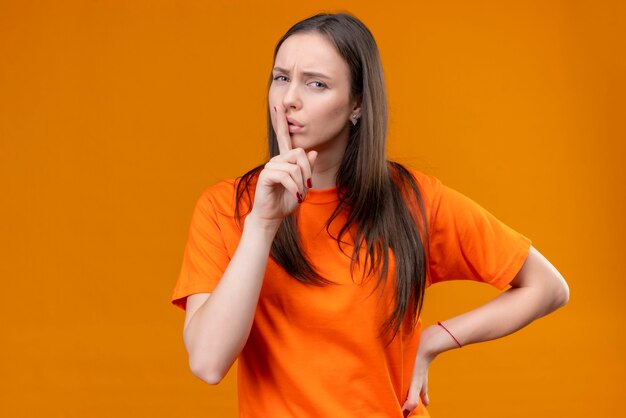 Young beautiful girl wearing orange t-shirt making silence gesture with finger on lips looking at camera with frowning face standing over isolated orange background