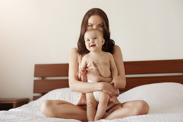 Young beautiful happy mother in sleepwear and her newborn baby sitting on bed in morning smiling playing together