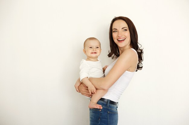 Young beautiful happy mother smiling laughing holding her baby daughter over white wall.