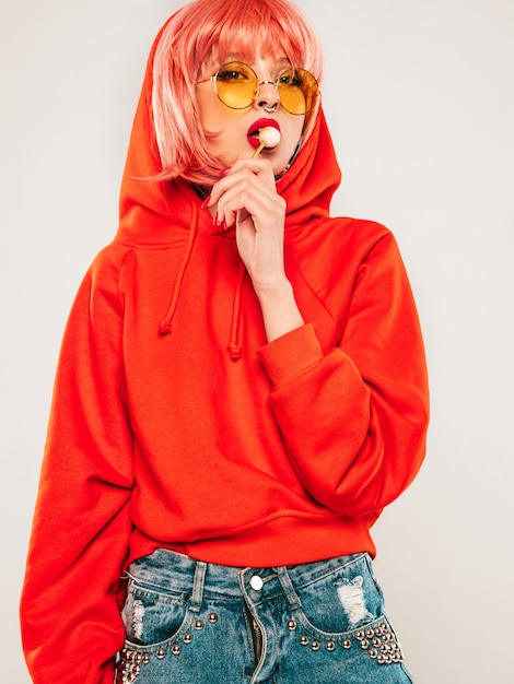 Free photo young beautiful hipster bad girl in trendy red summer red hoodie and earring in her nose.sexy carefree woman posing in studio on gray background in wig.hot model licking round sugar candy