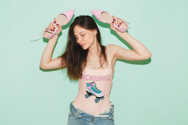 Young beautiful hipster woman posing against blue wall, holding pair of trendy shoes in hands