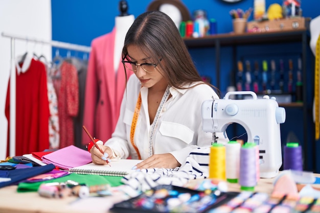 Young beautiful hispanic woman tailor drawing on notebook at clothing factory