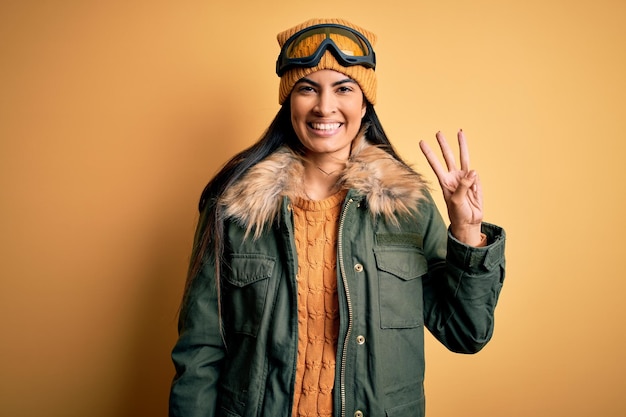 Free photo young beautiful hispanic woman wearing ski glasses and coat for winter weather showing and pointing up with fingers number three while smiling confident and happy