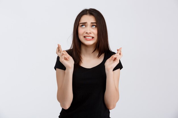 Young beautiful hopeful girl in black t-shirt posing with crossed fingers  over white wall