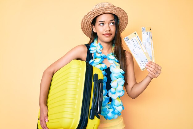 Young beautiful latin girl wearing summer hat and hawaiian lei holding cabin bag and boarding pass smiling looking to the side and staring away thinking.