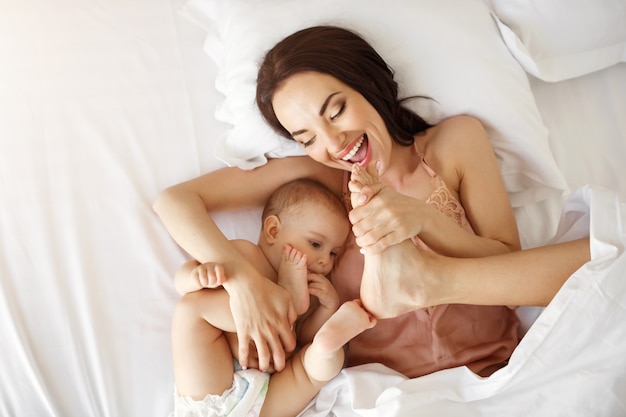 Young beautiful mom and newborn baby lying in bed smiling fooling at home. From above.