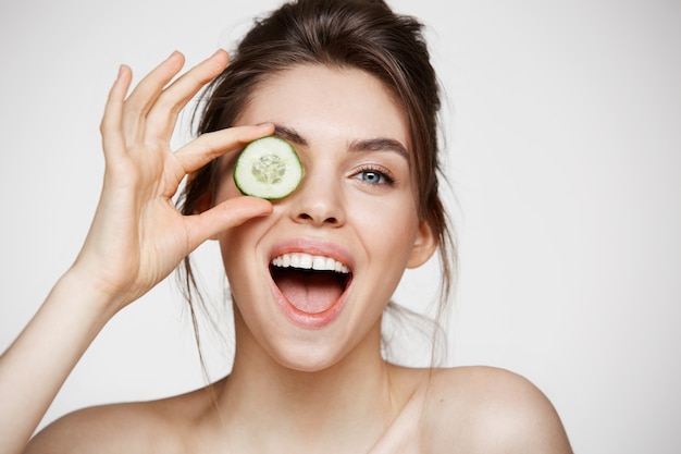 Free photo young beautiful naked girl smiling hiding eye behind cucumber slice looking at camera over white background.