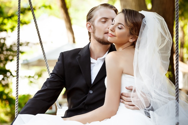 Free photo young beautiful newlyweds smiling, kissing, sitting on swing in park.