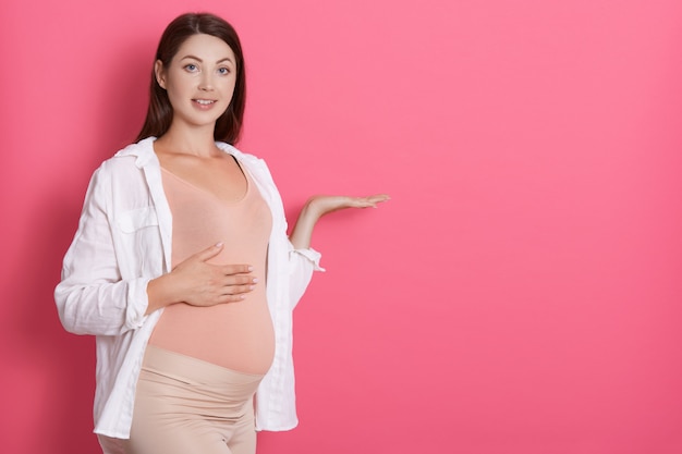 Young beautiful pregnant girl expecting baby isolated over pink space pointing aside with hands open palms showing copy space, presenting advertisement, looks smiling at camera.