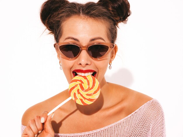 Young beautiful sexy hipster woman with red lips in sunglasses.Trendy girl in summer swimwear clothes. Eating candy lollipop