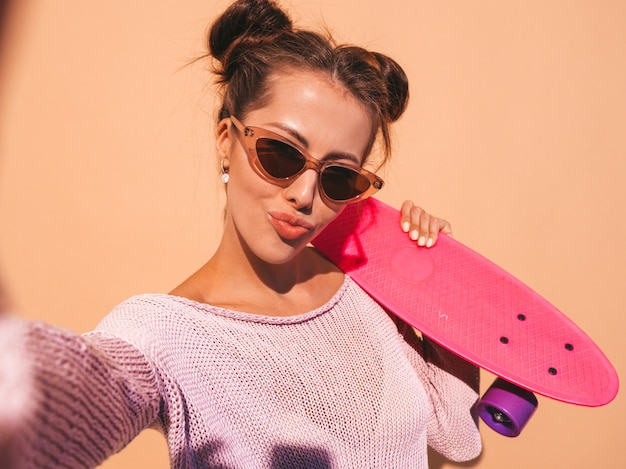 Free photo young beautiful sexy smiling hipster woman in sunglasses.trendy girl in summer knitted cardigan.female with pink penny skateboard,isolated on beige wall.taking selfie self portrait photos on phon