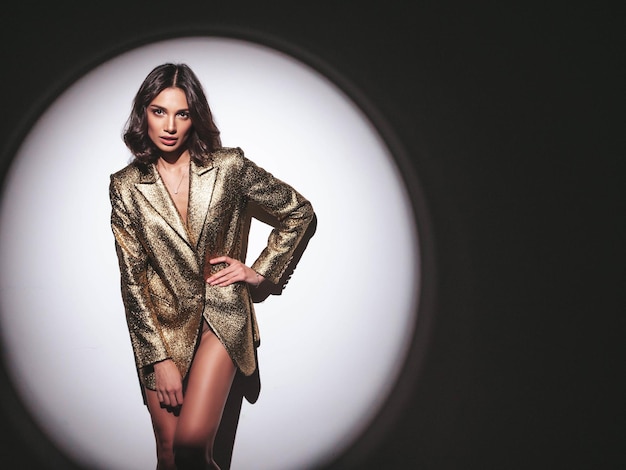 Young beautiful smiling brunette female in trendy evening gold jacket Sexy carefree woman posing near white wall in studio in a circle of lightFashionable model with bright makeup