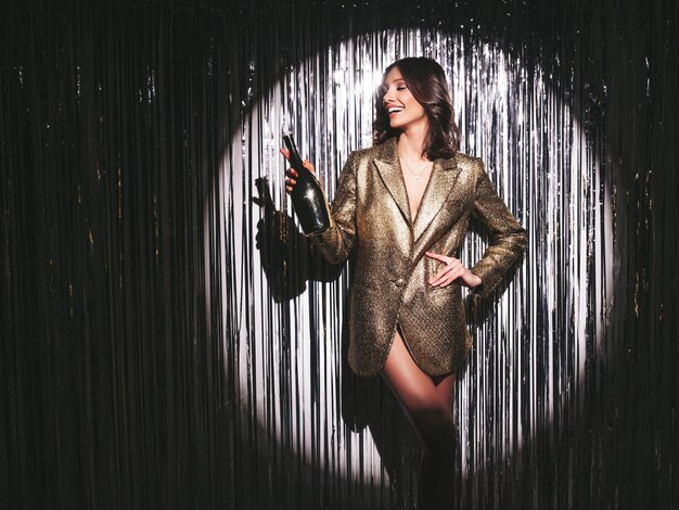 Young beautiful smiling brunette female in trendy evening golden jacketSexy carefree woman posing near silver shiny tinsel wall in studioFashionable model with bright makeup holding champagne bottle