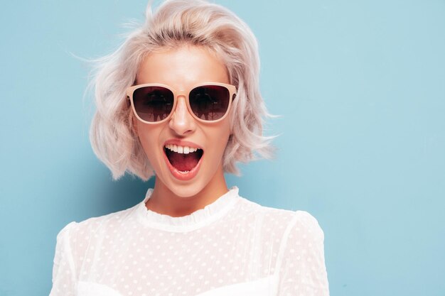 Young beautiful smiling female in trendy summer clothes Sexy carefree woman posing near blue wall in studio Positive blond model having fun in sunglasses Cheerful and happy Screaming and shouting