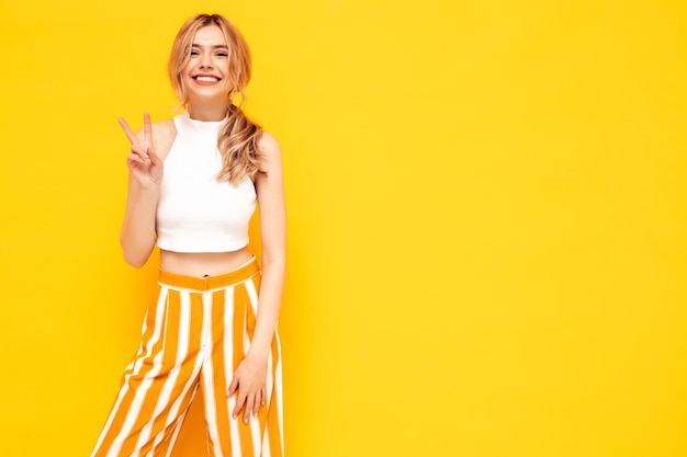 Free photo young beautiful smiling female in trendy summer clothes sexy carefree woman posing near blue yellow in studio positive model having fun cheerful and happy shows peace sign