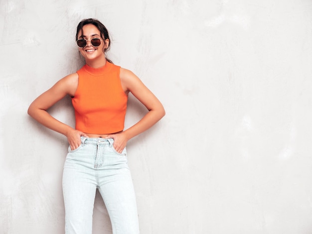 Free photo young beautiful smiling female in trendy summer clothes sexy carefree woman posing near grey wall in studio positive brunette model having fun and going crazy cheerful and happy in sunglasses