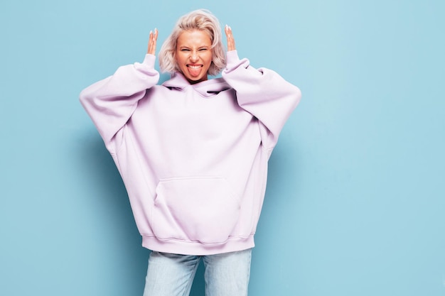 Young beautiful smiling female in trendy summer hoodie Sexy carefree woman posing near blue wall in studio Positive blond model having fun and going crazy Cheerful and happy